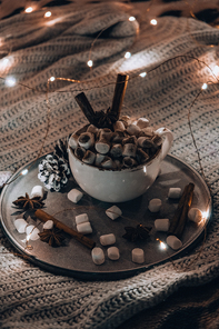 Cup with hot winter cacao and marshmallows on bed. Christmas lights. Pine cones decoration. Cozy winter days. Hygge
