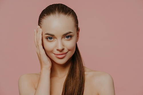 Isolated shot of pretty woman with combed pony tail, enjoys freshness of skin, looks with charming expression, stands bare shoulders, isolated on pink wall. Health care, makeup, spa and face lifting