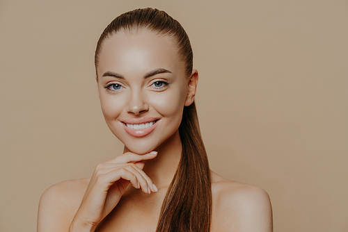 Isolated shot of young woman with European appearance has pure healthy smooth skin after doing daily cleansing procedures, smiles toothily, stands with bare shoulders indoor over beige wall.