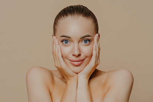 Headshot of young woman with surprised expression, touches face with both hands, has combed hair, natural makeup, healthy soft skin, stands against beige background. Facial treatment, cosmetology, spa