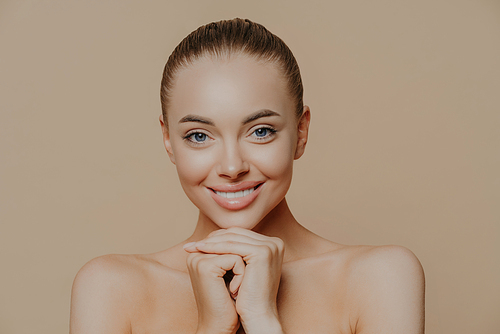 Close up shot of adult woman with fresh daily makeup, smiles toothily and keeps hands under chin, stands half naked indoor, feels refreshed after cosmetic procedures. Beauty and face care concept