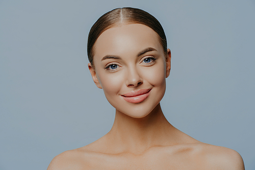 Headshot of attractive woman has beautiful facial features, natural makeup, dark combed hair, naked body, full lips, isolated on blue background, undergoes beauty treatments. Skin care concept