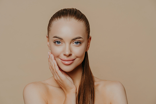 Photo of lovely young woman has smooth healthy skin after beauty procedures and receiving facial massage, stands bare shoulders, has long dark straight hair, isolated over beige wall cares of her body