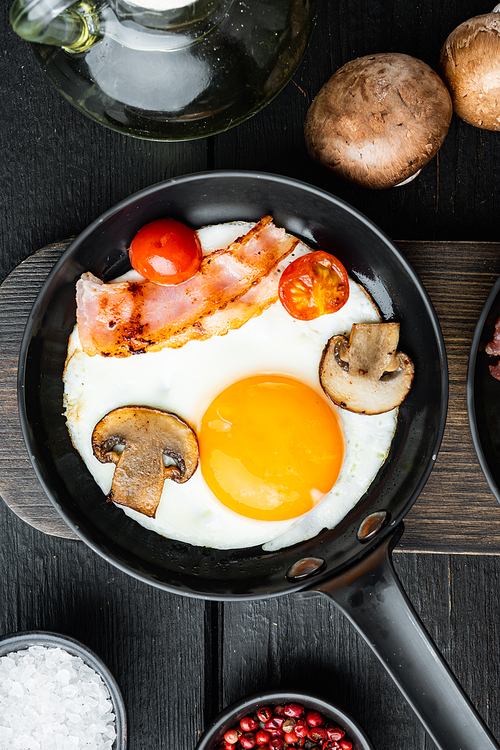 Fried eggs with cherry tomatoes and bread for breakfast in cast iron frying pan, on black wooden table background, top view flat lay