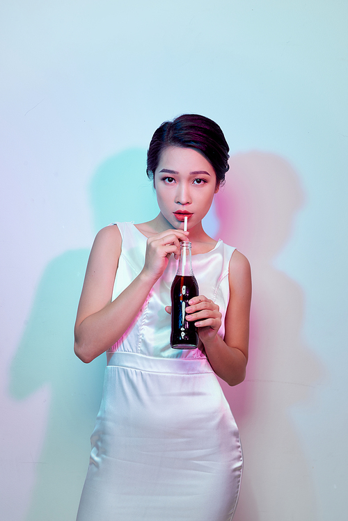 A beautiful tanned girl in a white silk dress and with red lipstick drinks a red drink from a glass bottle through a straw.