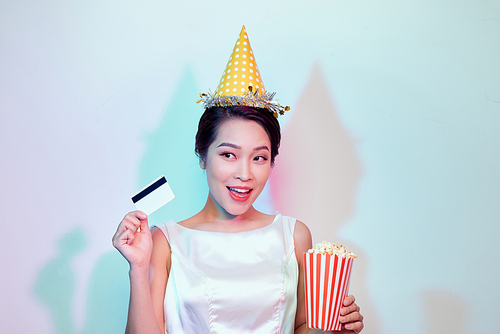 Portrait of young overjoyed attractive woman in white dress watching movie film, holding bucket of popcorn and credit card isolated on white background. Emotions in cinema concept