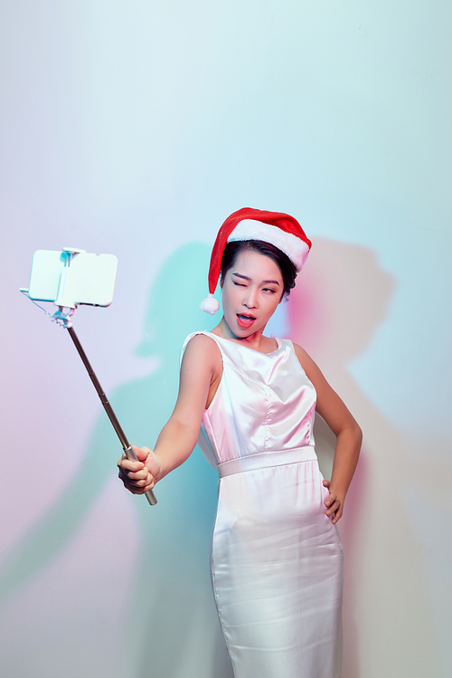 Modern holiday concept. Blonde woman in Santa hat taking Christmas picture of herself, selfie with smartphone on stick.