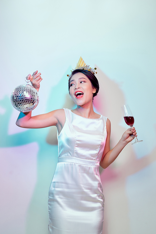 Portrait of a happy beautiful woman in white dress having a party and drinking champagne while standing with disco ball isolated over light background