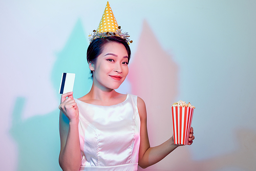 Portrait of young overjoyed attractive woman in white dress watching movie film, holding bucket of popcorn and credit card isolated on white background. Emotions in cinema concept