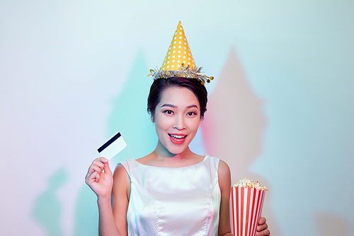 Portrait of young overjoyed attractive woman in white dress watching movie film, holding bucket of popcorn and credit card isolated on white background. Emotions in cinema concept