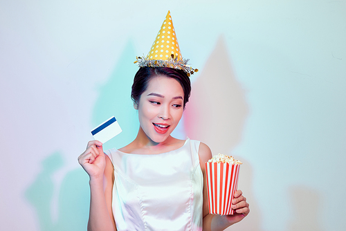 Portrait of young overjoyed attractive woman in white dress watching movie film, holding bucket of popcorn and credit card isolated on white background. Emotions in cinema concept