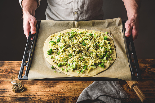Man holding baking dish with uncooked pizza with broccoli, pesto sauce, spices and cheese