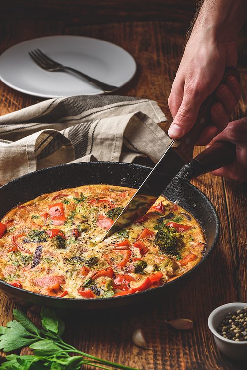 Hand with knife slices vegetable frittata with broccoli, red bell pepper and red onion in cast iron pan