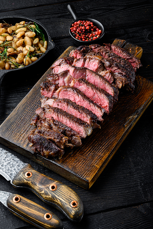 Fresh grilled meat. Grilled beef steak medium rare black angus rib eye steak set, on wooden serving board, with white beans and rosemary in cast iron pan, on black wooden table background