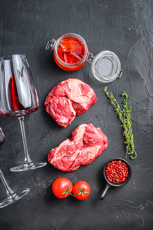 Raw chuck eye roll steak cuts, with red wine glass , rosemary, spicy chili oil on black textured background  top view