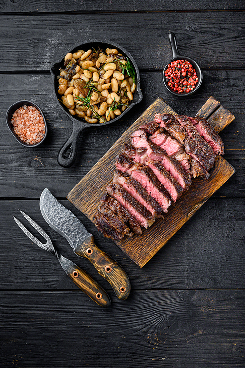 Sliced grilled meat steak Rib eye medium rare set, on wooden serving board, with white beans and rosemary in cast iron pan, with meat knife and fork, on black wooden table background, top view flat lay, with copy space for text