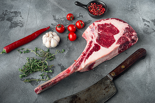 Dry aged raw tomahawk beef steak with ingredients for grilling set, and old butcher cleaver knife, with seasoning and herbs, on gray stone background