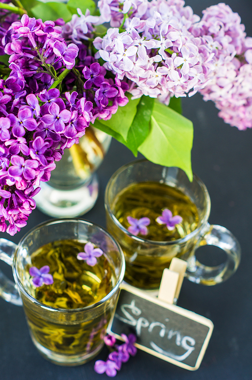 Cup of green tea and bright purple lilac flowers on a rustic wooden table with note