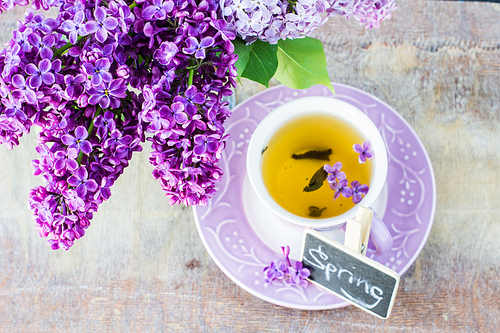 Cup of green tea and bright purple lilac flowers on a rustic wooden table with note