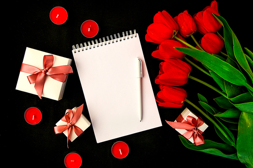 Red tulips, blank card or notepad with pen and candles, gift box isolated on black background, copy space for text, holiday concept, springtime
