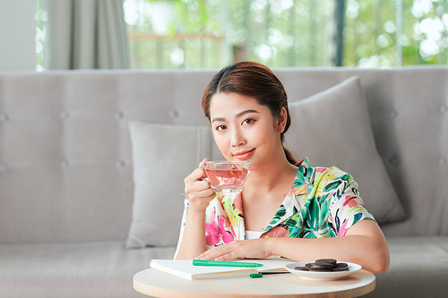 Young cheerful woman sitting on floor writing in diary and drinking morning tea