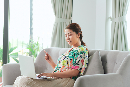 Beautiful young woman working on laptop computer while sitting at the living room, showing credit card