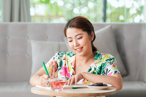 Woman writing notes in an agenda or diary sitting on a couch in the living room at home