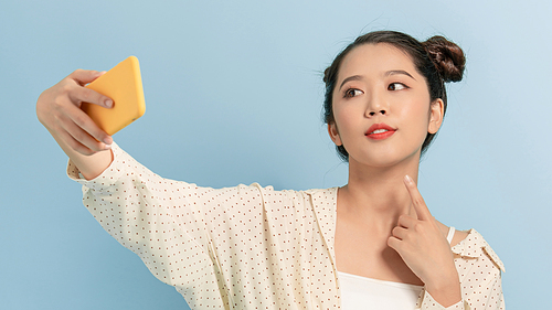 Smiling adorable Asian female taking selfie photo on smartphone with positive expression over blue background.