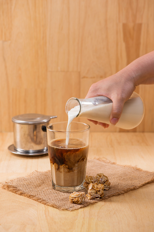 Pouring milk in to glass of coffee on a wooden table.