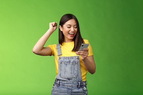 Triumphing cute joyful asian girl hold smartphone, fist pump celebrating good news, read good news message, look telephone screen smiling happily, achieve success got persmission party.