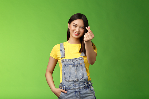 Lovely tender carefree asian girlfriend show korean love sign, make finger heart smiling cute, showing passion and affection, stand green background in overalls and yellow t-shirt, express sympathy.