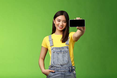 Attractive cheerful asian brunette female introduce smartphone game, hold telephone horizontal, show cellphone display, smiling delighted, give recommendation what app download, green background.