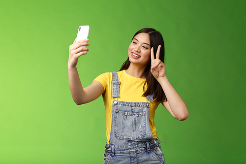Tender cute asian brunette female, hold smartphone, posing selfie, look telephone screen, photographing, show victory peace sign, joyfully chatting friend abroad, stand green background. Copy space