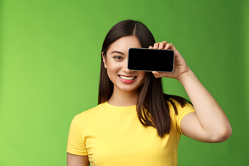 Close-up cheerful optimstic asian woman promote smartphone app hold telephone near eye gaze camera satisfied, smiling broadly introduce game, cool new application, stand green background.