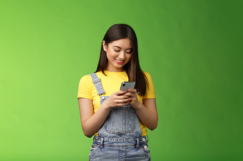 Outgoing cute asian brunette woman scroll online shop, purchase summer tour internet, hold smartphone, look amused telephone screen, use app, edit photos post social media, stand green background.