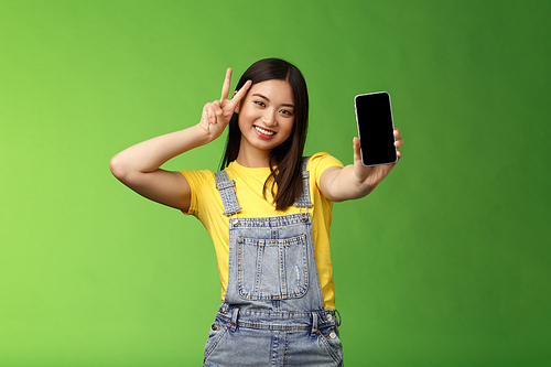 Cute carefree asian brunette showing application on smartphone screen, make victory peace sign, smiling joyfully, brag social media popularity, followers amount, stand green background.
