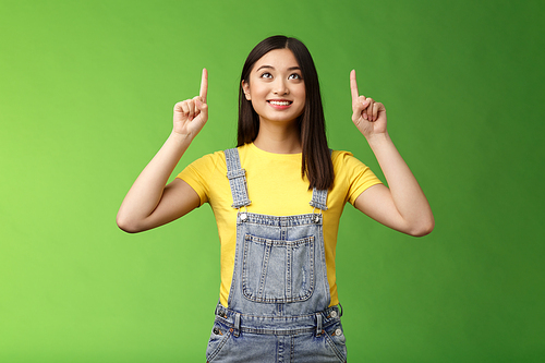 Dreamy hopeful cute asian girl look pointing up amused, smiling delighted, contemplate interesting object, enjoy stargazing, grinning satisfied, gazing top promo, stand green background.