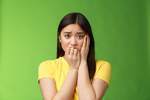 Innocent insecure timid asian scared girl panicking, standing afraid victim terrified, touch cheek shocked, frowning stunned, biting fingernails, anxiously stare camera, green background.