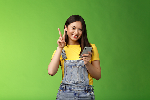 Cute tender asian woman hold smartphone, show victory peace sign, look camera delighted, carefree pose near green background, win online giveaway, triumphing joyfully beat score game.