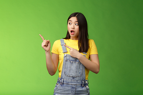 Shocked gasping alarmed pretty asian girl drop jaw impressed, turn pointing left hear loud bang, stand astonished speechless, gazing curious event, pose green background. Emotions concept