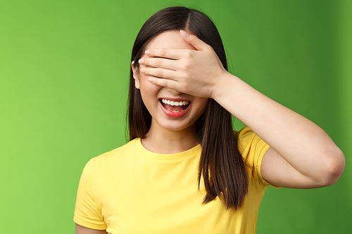 Lively joyful friendly-looking asian girl shut eyes palm, laughing happily, standing amused anticiapting b-day gift, counting ten, playing hide-n-seek, stand green background entertained.