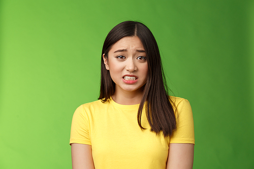 Anxious worried cute asian girl brunette clench teeth frowning shaking from fear look guilty afraid, stand nervous awaiting punishment, green background, apologizing ashamed. Copy space