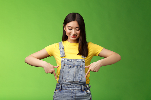 Studio shot easygoing cute asian female introduce new promo, pointing looking down amused pleasantly smiling, gladly show interesting thing, stand green background happy excited. Copy space