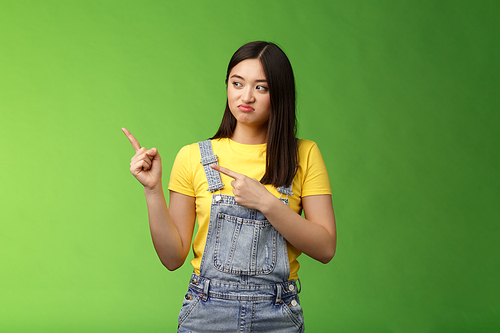 Upset cute asian female student pulling face regret shame, pity missing good chance buy product sale, pointing looking left disappointed, jealously gaze coworker, stand green background.