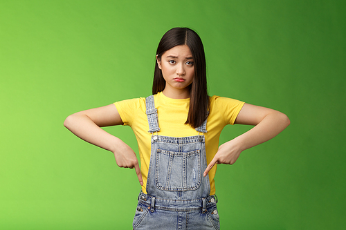 Sad asian girl in dispair feel upset, frowning sulking pessimistic, pointing down look camera pulling sorrow uneasy face, express sadness and frustration, stand green background. Copy space