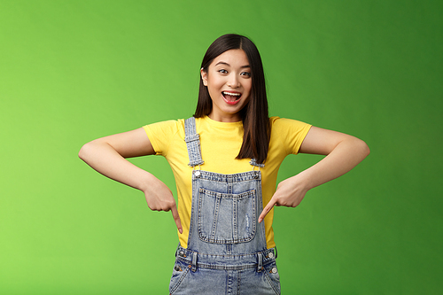 Lively energized asian cute female promote advertisement pointing fingers down, show you cool place, smiling camera joyful, telling great pleasant news stand green background emotional.
