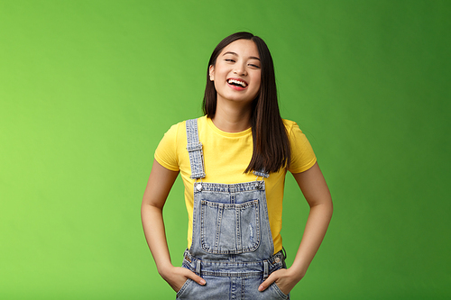 Carefree enthsuastic outgoing attractive teenage asian girl having fun laughing friends, hold hands overalls pockets smiling toothy, take party easygoing conversation, stay positive green background.