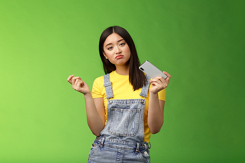 Bored indifferent cool urban asian girl waste time summer holiday alone, playing smartphone game, interrupt texting, browsing internet, gesturing reluctant, look sad, losing battle, green background.