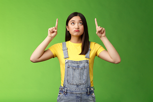 Silly upset cute asian girl look pointing up sad, gloomy watching top side, grimacing displeased, dreaming have new product, regret not have money effort cool thing, stand green background.