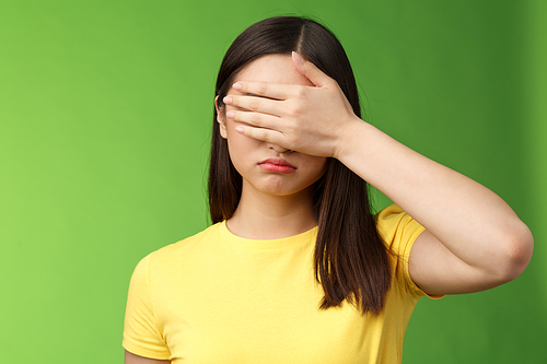 Sad tired asian girl dark haircut, shut eyes hold hand sight, grimacing, feeling gloomy and upset, stand green background depressed, unwilling watch how life ruins, pose yellow t-shirt.